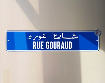 Puede incluir: Un letrero de calle rectangular azul con el texto blanco "RUE GOURAUD" en inglés y árabe. El letrero está montado en una pared de color claro. El sitio web "www.whynatt.com" está en la parte inferior.