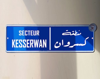Puede incluir: Un letrero azul y blanco con el texto "SECTEUR KESSERWAN" en francés y "كسروان" en árabe. El letrero está montado en una pared.