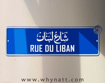 Puede incluir: Un letrero de calle azul con letras blancas que dice "Rue du Liban" en francés y "شارع لبنان" en árabe. El letrero está montado en una pared.