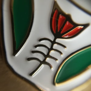 May include: A white enamel pin with a gold outline. The pin features a stylized red flower with a gold stem and leaves. The flower has a green center.