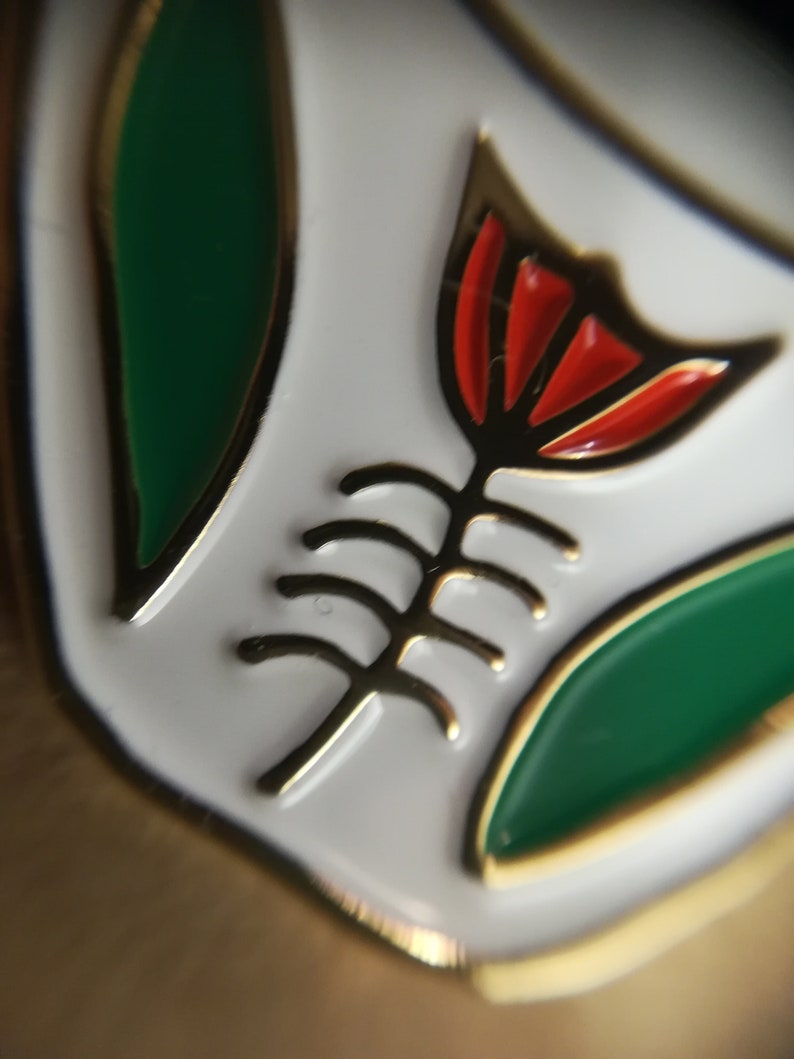 May include: A white enamel pin with a gold outline. The pin features a stylized red flower with a gold stem and leaves. The flower has a green center.