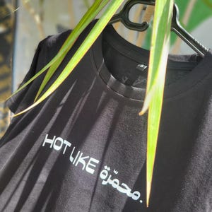 May include: Black t-shirt with the white text "HOT LIKE" and Arabic script. The shirt is hanging on a black hanger, with green and yellow leaves in the background.