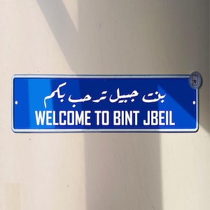 Puede incluir: Un letrero azul y blanco con el texto "Bienvenido a Bint Jbeil" en inglés y árabe. El letrero está montado en una pared.