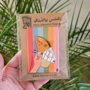 May include: A colorful magnet with a cartoon illustration of a hand holding a Lebanese manakish, a flatbread with toppings, wrapped in a white paper bag. The bag has the text "Angus" written on it. The magnet has a rainbow striped background and the text "Very Lebanese Magnets" and "WWW.WHYATT.COM".