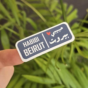 Può includere: Un adesivo bianco con testo blu e rosso. L'adesivo dice "Habibi Beirut" in inglese e "بيروت" in arabo con un simbolo di cuore rosso.