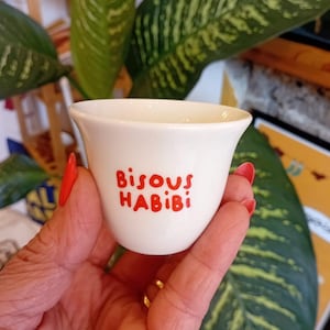 Puede incluir: Una pequeña taza de cerámica blanca con las palabras "Bisous HABIBI" en letras rojas. La taza se sostiene en una mano con esmalte de uñas rojo, con plantas verdes de hojas en el fondo.