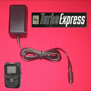 Könnte beinhalten: Schwarze Turbo Express Handheld-Konsole mit schwarzem Netzteil und Kabel. Die Konsole hat einen kleinen Bildschirm und Tasten. Auf dem Netzteil steht "Turbo Express".