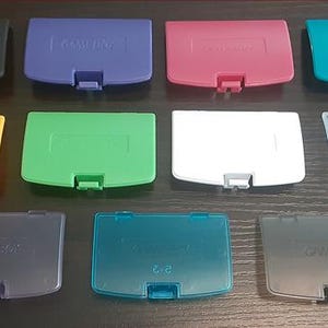 May include: A collection of ten Game Boy cartridge covers in various colors, including black, blue, pink, teal, yellow, green, white, and translucent. Each cover has a rectangular shape with a small latch.