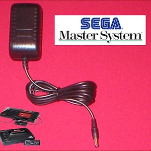 May include: Black power adapter for a Sega Master System console, with the Sega Master System logo in white and green. The adapter has a black cord and a DC connector. Two Sega Master System consoles are shown in the image.