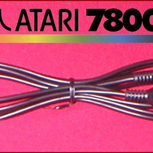 以下が含まれることがあります： RCAとBNCコネクタを備えた黒いAtari 7800ビデオゲームケーブル。ケーブルは丸められ、黒いタイで固定されています。Atariのロゴと「ATARI 7800」の文字が赤い背景の上にあります。