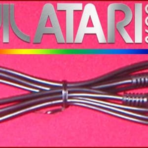 Puede incluir: Cable de vídeo negro Atari 2600 con dos conectores. El logotipo de Atari y "2600" se muestran encima del cable sobre un fondo rojo. Una franja de color arcoíris está debajo del logotipo.