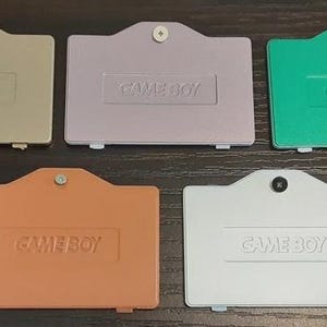 Puede incluir: Cinco tapas de batería de Game Boy en varios colores: beige, lavanda, verde, naranja y blanco. Cada tapa tiene la palabra "GAME BOY" en relieve. Las tapas son rectangulares con una parte superior curvada y un tornillo en el centro.