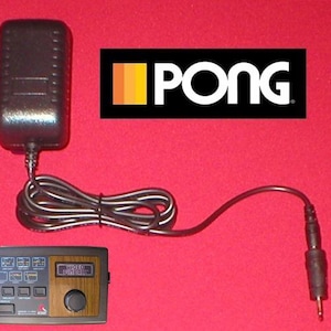Peut inclure: Une console de jeu vidéo Atari Pong avec un adaptateur secteur et un câble noirs. La console a un panneau imitation bois avec des boutons et un cadran. Le logo Pong est affiché en haut à droite.