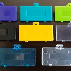 Puede incluir: Una colección de ocho tapas de batería para Game Boy Advance en varios colores, incluyendo morado, turquesa, verde, negro, amarillo y opciones translúcidas. Cada tapa tiene forma rectangular con un cierre.