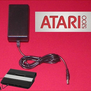 May include: A black Atari 5200 console with a controller and power supply. The power supply is black and rectangular. The Atari 5200 logo is visible in red on a white background.
