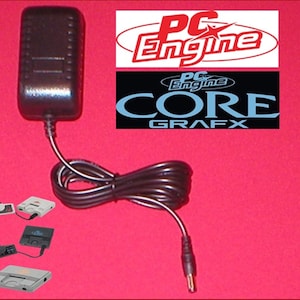 Puede incluir: Adaptador de corriente negro con cable y conector negros. La imagen incluye el texto "PC Engine" en rojo y blanco, y "PC Engine Core Grafx" en azul y blanco. Se muestran varias consolas de juegos en la esquina inferior izquierda.