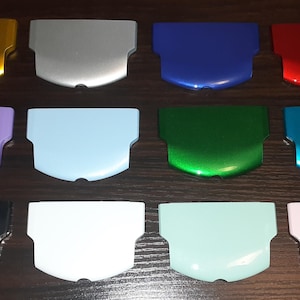 Może przedstawiać: Kolekcja dwunastu błyszczących plastikowych osłon w różnych kolorach, w tym żółtym, srebrnym, niebieskim, czerwonym, fioletowym, jasnoniebieskim, zielonym, turkusowym, czarnym, białym, miętowym i różowym. Osłony mają zakrzywioną górę i prostokątną podstawę.