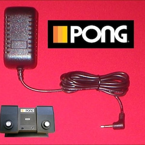 May include: An Atari Pong console with a black power adapter and cable. The console has two control knobs and the word "PONG" is printed on the console and on a black rectangle with a colourful stripe design.