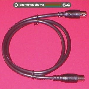 May include: A black Commodore 64 video cable with two connectors. The cable is looped and has the Commodore 64 logo in the upper left corner and the number 64 in the upper right corner. The background is a solid pink color.