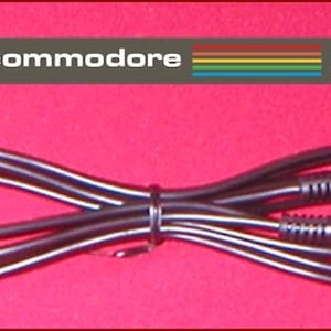 May include: Black video cable with RCA and BNC connectors, for a Commodore 64. The cable is looped and secured with a black tie. The Commodore 64 logo and text are in the upper left corner, with a rainbow stripe design.