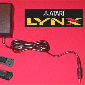 Puede incluir: Un cargador de consola de juegos portátil Atari Lynx. El cargador y el cable negros están conectados a una consola de juegos portátil negra y gris. El logotipo de Atari Lynx se muestra sobre un fondo rojo.