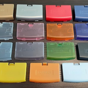 May include: A selection of Game Boy cartridge covers in a range of colours, such as gold, red, blue, and green. Some covers are translucent, and all feature the "GAME BOY" logo. The covers are rectangular with a small tab at the top.