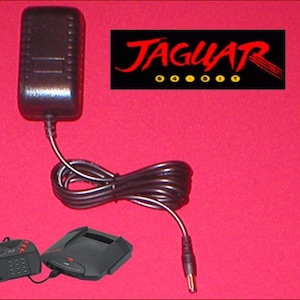 May include: Black power adapter with a black cable, a black game console, and a black controller on a red background. The text "JAGUAR" is displayed in red with yellow dots.