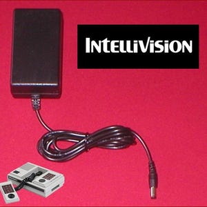 May include: A black power adapter with a black cord, connected to a vintage Intellivision game console and controller. The Intellivision logo is displayed on a black rectangle. The background is red.