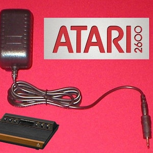 Op de afbeelding: Een Atari 2600 videogameconsole met een voedingsadapter en kabel. De console is zwart met een houtnerf bodem en zilveren accenten. Het Atari 2600 logo wordt weergegeven op een witte achtergrond met rode tekst.