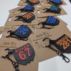 May include: A collection of black keychains with various names and numbers in orange, blue, and white. Each keychain has a metal chain and clasp, and is attached to a brown card with the text "AGS Customizations".