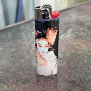 May include: A lighter with a personalised photo of a couple. The woman is wearing a face mask and a white shirt. The man is wearing a black shirt. The lighter is silver with a red top.