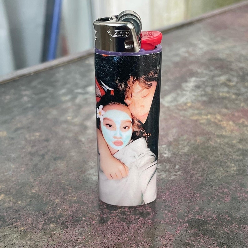 May include: A lighter with a personalised photo of a couple. The woman is wearing a face mask and a white shirt. The man is wearing a black shirt. The lighter is silver with a red top.