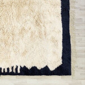 May include: A cream-coloured, shaggy rug with a dark navy border and fringe. The rug has a textured, plush appearance, with a contrasting dark navy border that frames the cream-coloured centre. The fringe adds a decorative element.