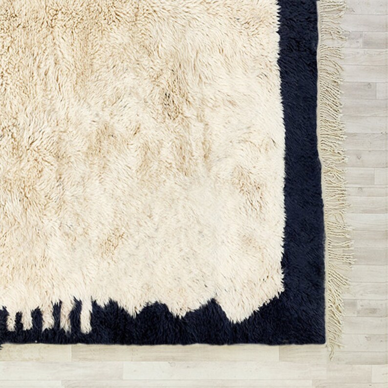 May include: A cream-coloured, shaggy rug with a dark navy border and fringe. The rug has a textured, plush appearance, with a contrasting dark navy border that frames the cream-coloured centre. The fringe adds a decorative element.