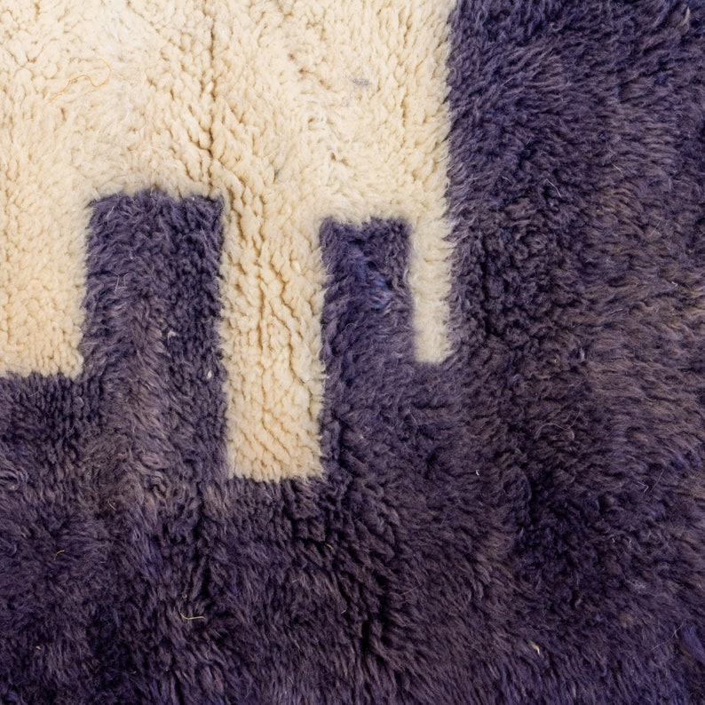 May include: Close-up of a textured rug with a geometric design. The rug has a deep purple background and a cream-coloured pattern. The design appears to be a series of connected shapes, creating a modern aesthetic.