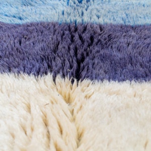 May include: Close-up of a textured rug with horizontal stripes. The rug features bands of light blue, deep purple, and off-white. The fibres are thick and create a soft, plush appearance. The rug's design is simple and modern.