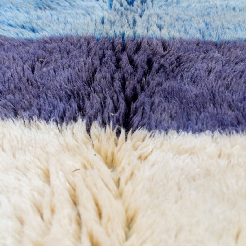 May include: Close-up of a textured rug with horizontal stripes. The rug features bands of light blue, deep purple, and off-white. The fibres are thick and create a soft, plush appearance. The rug's design is simple and modern.