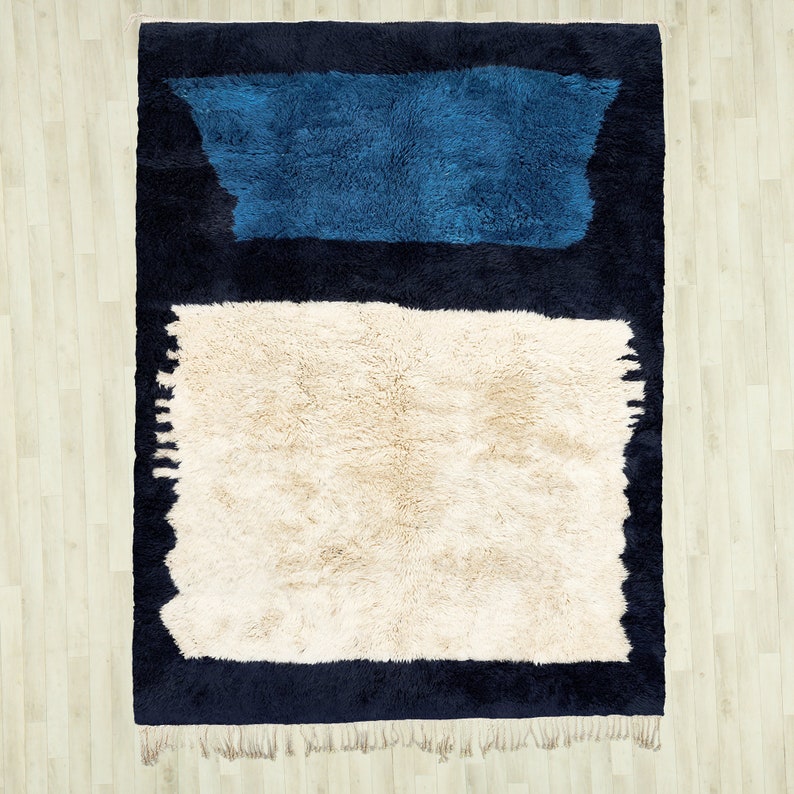 May include: A rectangular rug with a cream-coloured centre and a dark blue border. A blue rectangular shape is at the top of the rug. The rug has a shaggy texture and fringe along the bottom edge. The rug is on a light wood floor.