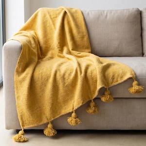 Puede incluir: Una manta de color amarillo mostaza con borlas decorativas sobre un sofá beige. La manta tiene un aspecto texturizado y está doblada para mostrar las borlas a lo largo del borde. Las borlas son de color amarillo mostaza a juego.