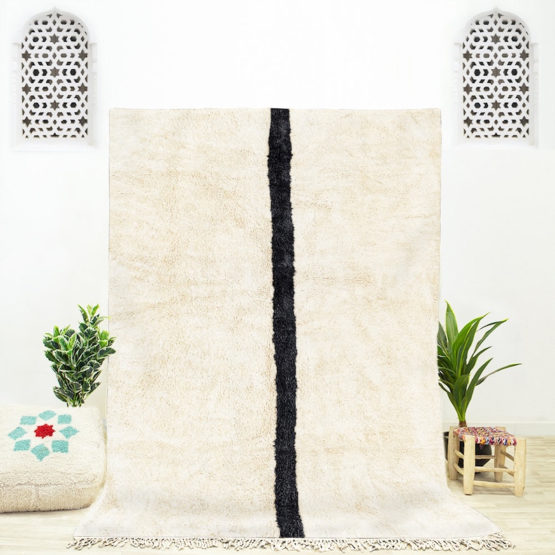 Puede incluir: Una alfombra de pelo blanco con una sola raya negra en el centro. La alfombra tiene un borde de flecos y se muestra en una habitaci&oacute;n con paredes blancas y dos ventanas decorativas.