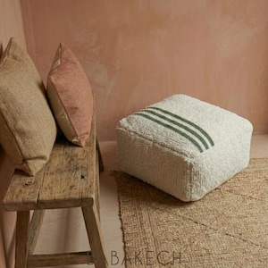 Può includere: Una panca rustica in legno sostiene due cuscini, uno beige e uno rosa. Un pouf quadrato bianco e strutturato con tre strisce verdi poggia su un tappeto tessuto. Lo sfondo è un muro rosa.