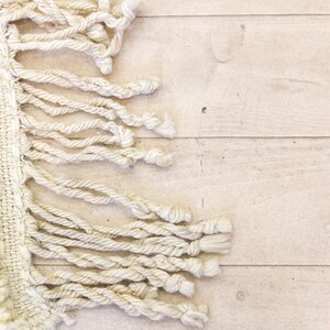 May include: Close-up of a cream-coloured woven textile with long, knotted fringe. The textile is placed on a light-coloured wooden surface. The fringe hangs down from the edge of the woven material.