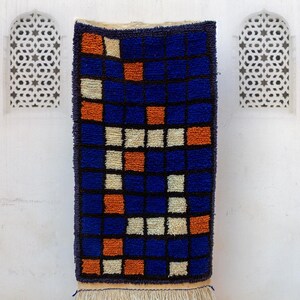 May include: A rectangular, hand-woven rug with a geometric pattern. The rug features a grid of blue squares with orange and off-white accents. The rug has a fringe at the bottom and is displayed against a white wall.