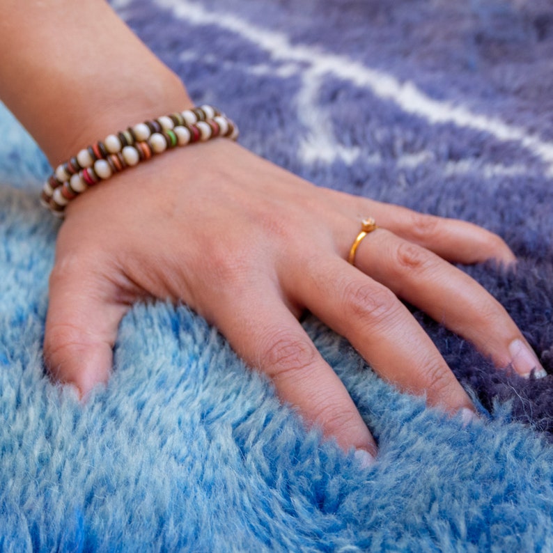 May include: A hand rests on a blue and white rug, with a beaded bracelet and a gold ring. The bracelet features alternating white, brown, and green beads. The rug has a fluffy texture and a gradient of blue hues.