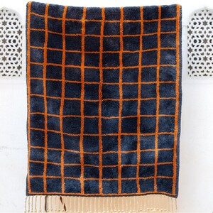 May include: A navy blue towel with a grid pattern in orange. The towel has a fringed edge at the bottom. The grid pattern is evenly spaced across the towel's surface. The towel is hanging against a white wall.