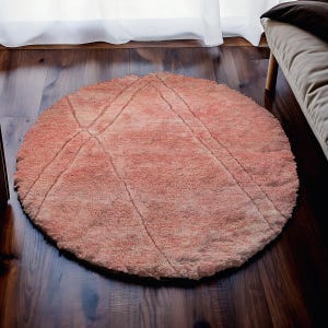 May include: A round, peach-colored rug with a geometric design. The rug features a soft, textured surface and is placed on a dark wood floor. The design includes diagonal lines that intersect in the center.