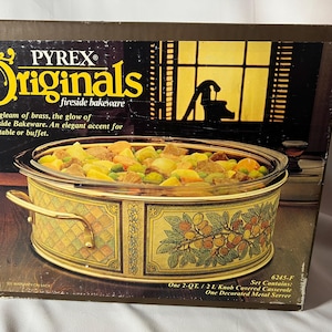 May include: Pyrex Originals fireside bakeware set. The box features a casserole dish filled with food. The dish has a gold-colored metal base with a decorative floral design. The box has text that reads "The gleam of brass, the glow of Fireside Bakeware."