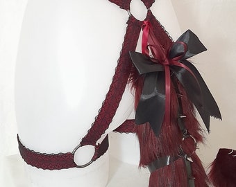 May include: A red and black lace harness with a faux fur tail and a black bow. The harness has two metal rings and the tail has spikes.