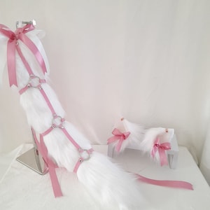 Peut inclure: Une queue en fausse fourrure blanche et un ensemble d'oreilles assorties avec des rubans roses. La queue est dotée d'un harnais avec des anneaux argentés et des sangles roses. Les oreilles sont fixées à de petites plateformes blanches, chacune ornée d'un nœud rose.