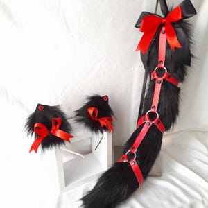 Op de afbeelding: Een zwarte faux fur staart- en orenset met rode accenten. De staart heeft een rood harnas en strik, terwijl de oren rode strikken en hartdetails hebben. De set is ontworpen voor cosplay of kostuumgebruik.